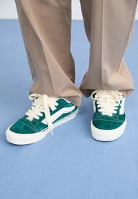 Zapatillas de ante verde con cordones y acentos blancos, que presentan un perfil bajo y una punta redondeada. Combinadas con pantalones beige sobre un fondo azul.