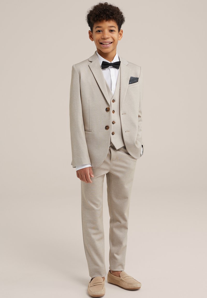 Traje de tres piezas beige con botones marrones, camisa blanca, pajarita negra y un pañuelo de bolsillo. Se lleva con mocasines beige. Textura de tela suave.