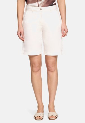 Kobieta w białych szortach do kolan z przednimi kieszeniami i beżowych sandałach wsuwanych, stojąca na jednolitym tle.