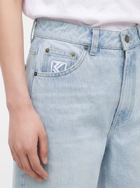 Jean en denim bleu clair avec un gros plan sur la poche avant et une main reposant à côté, portant une chemise blanche rentrée.