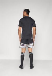 Athletisches Ensemble mit einem eng anliegenden schwarzen Shirt, glänzenden, schimmernden Shorts mit goldenen Akzenten, knielangen schwarzen Socken und schwarzen Fußballschuhen.