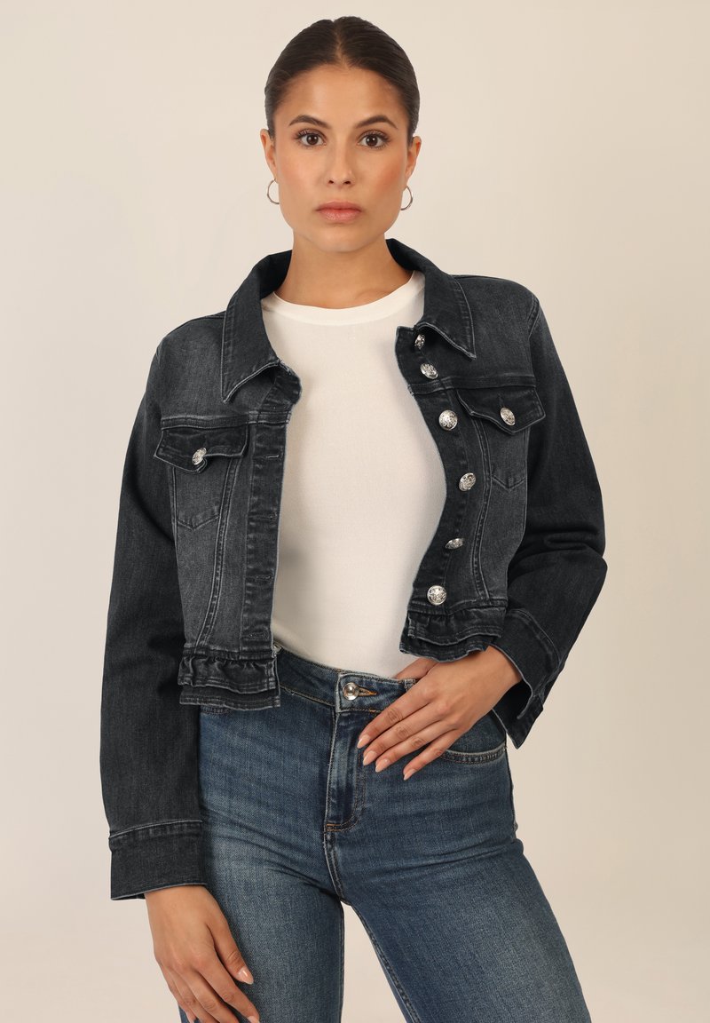 Dark denim jacket with a structured design, silver button accents, and a frayed hem. Worn over a white shirt and paired with blue jeans.