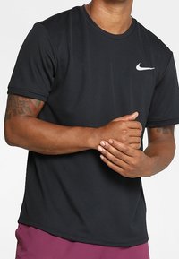 Muž v černém tričku s krátkým rukávem Nike a fialových šortkách, s rukama spojenýma před trupem, s viditelnými tetovanými pažemi.