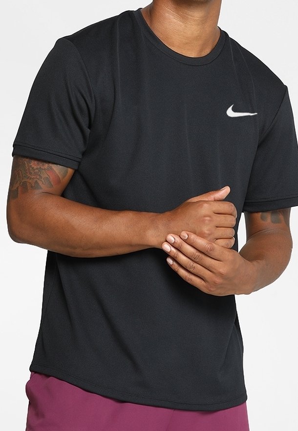 Muž v černém tričku s krátkým rukávem Nike a fialových šortkách, s rukama spojenýma před trupem, s viditelnými tetovanými pažemi.