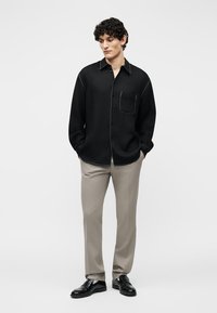 Homme debout les mains dans les poches, portant une chemise noire à boutons avec des coutures blanches, un pantalon beige et des chaussures habillées noires sur fond uni.