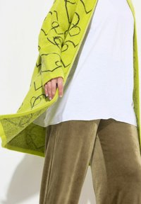Cardigan verde a fantasia con una texture a maglia larga, abbinato a una camicia bianca e pantaloni di velluto verde oliva lucidi, che mostrano un look casual a strati.