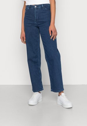 Vrouw draagt een donkerblauwe jeans met hoge taille en rechte pijpen, gecombineerd met witte plateau sneakers, tegen een eenvoudige lichte achtergrond.