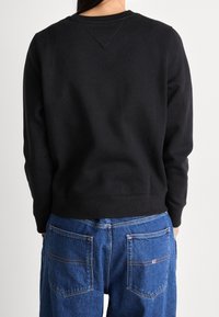 Felpa nera con scollatura rotonda e polsini a coste, abbinata a pantaloni in denim blu con tasche posteriori visibili e dettagli di cucitura.
