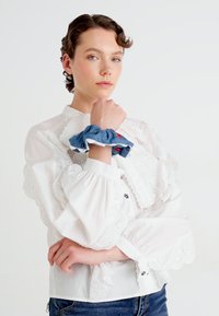 Mulher vestindo uma blusa de renda branca e um scrunchie de denim azul no pulso, posando com os braços cruzados contra um fundo branco.