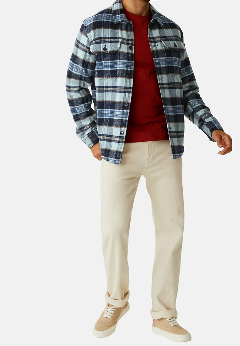 Camicia di flanella a quadri blu e bianca con due tasche sul petto, indossata sopra una t-shirt rossa. Abbinata a pantaloni color crema e sneakers marrone chiaro.