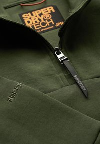 Sudadera de forro polar verde con cuello con cremallera, texto en relieve "SUPER" y una etiqueta prominente "SUPERDRY TECH" en naranja. Detalle del tirador de la cremallera en negro.