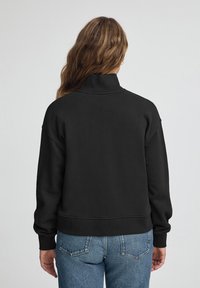 Czarna bluza z wysokim kołnierzem, prążkowanymi mankietami i luźnym fasonem, zestawiona z niebieskimi dżinsami o klasycznym pięciopocketowym kroju.