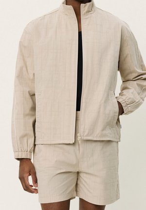 Mand iført en beige lynlåstrøje med elastiske manchetter og matchende beige shorts med subtilt ternet mønster.