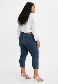 Jean skinny en denim bleu foncé avec un ourlet court, présentant un design classique de poche arrière et une étiquette en cuir marron sur la ceinture.