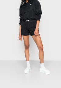 Giacca leggera nera a vento con mezzi zip, tasca frontale e polsini elastici, abbinata a pantaloni corti coordinati e sneaker bianche con zeppa.