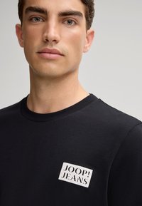 Zwarte ronde hals t-shirt van gladde stof, met een witte rechthoekige logopatch met de tekst "JOOP! JEANS" op de voorkant.