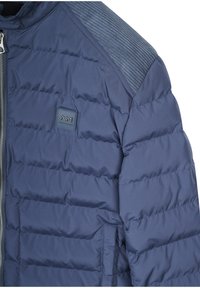 Mornarski puffer kaput od glatkog materijala, sa kviltiranim dijelovima, zatvaranjem na zip i rebrastim detaljima na ramenima. Mali logo na prsima.