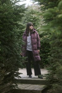 Lunga giacca a vento viola con cappuccio, indossata sopra una maglietta a righe, abbinata a pantaloni neri larghi. Circondata da alberi sempreverdi su un sentiero di legno.