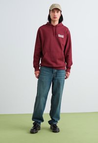Maroon hoodie with a front pocket and logo, paired with loose blue denim jeans and black shoes. Finished with a beige cap.