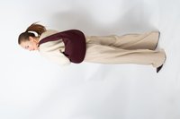 Femme en pull crème et pantalon large beige portant un grand sac à bandoulière tressé bordeaux, regardant sur le côté contre un fond blanc.