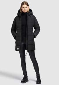 Sort puffjakke med hætte, med lynlås foran, quiltet mønster og sidelommer. Bæres over en sort turtleneck og leggings.