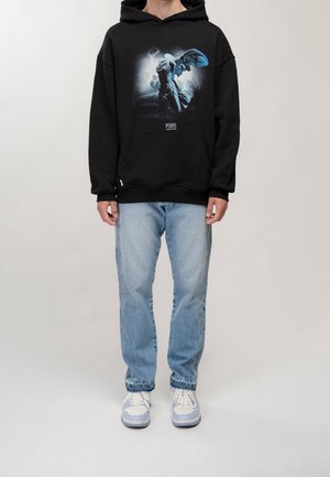 Sudadera negra con diseño gráfico azul de una figura, combinada con jeans azul claro y zapatillas blancas. La tela parece suave con un ajuste relajado.