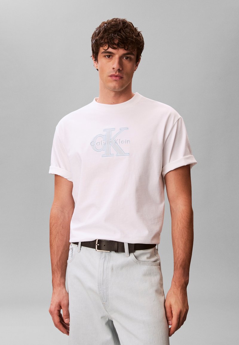 Νεαρός άνδρας που φοράει λευκό t-shirt Calvin Klein με ρολά μανίκια, τυλιγμένο μέσα σε ανοιχτόχρωμο τζιν παντελόνι  με μαύρη ζώνη, μπροστά σε ένα απλό γκρι φόντο.