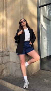 Blazer preto, T-shirt branca, calções de desporto azuis, ténis brancos e mala preta. De pé contra uma parede de pedra com uma janela moderna.