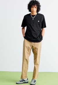 Black short-sleeve t-shirt with a logo on the chest, paired with beige chinos and gray-black sneakers with stripes. Curly hair visible.