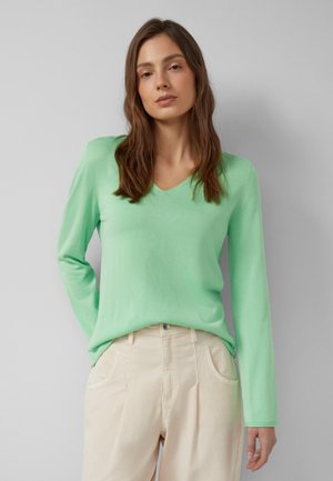 Vrouw met lang bruin haar, gekleed in een lichtgroene trui met V-hals en beige broek, staand tegen een effen grijze achtergrond.