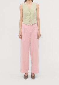 Kobieta w beżowej bezrękawnikowej kamizelce z guzikami, w szerokich różowych spodniach z wysokim stanem i brązowych spiczastych butach, stojąca na jednolitym tle.