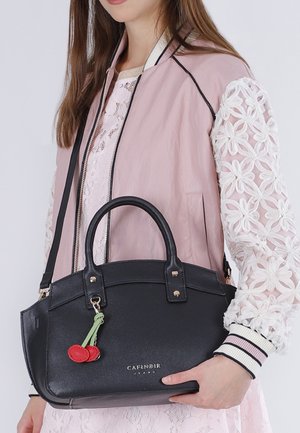 Mujer con chaqueta rosa con mangas de encaje floral sostiene un bolso de cuero negro con un adorno de cereza.
