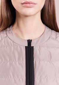 Lätt vadderad jacka i grått med hög krage, som har en central svart dragkedja och quiltad textur.