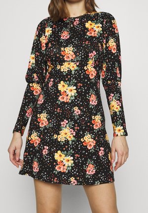 Frau, die ein schwarzes langärmeliges Kleid mit gelben, orangefarbenen und rosafarbenen Blumenmustern trägt. Das Kleid hat eine taillierte Taille und einen runden Ausschnitt.