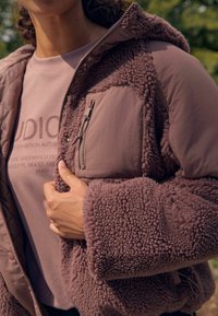 Zbliżenie na osobę w fioletowej polarowej kurtce z teksturowanym materiałem i zamykaną na zamek kieszenią na piersi, trzymającą kurtkę otwartą na zewnątrz.