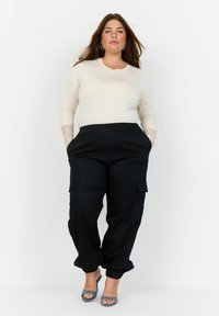 Helles beiges Langarmshirt mit Spitzenakzenten an den Handgelenken, kombiniert mit schwarzen Cargo-Hosen mit Seitentaschen und elastischen Bündchen. Blaue Schuhe.