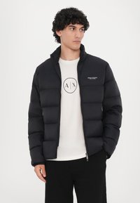 Sort puffer-jakke med stående krave, quiltet design og lynlås foran. Båret over en lysefarvet T-shirt med trykt logo.