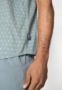 Ανοιχτό μπλε πουκάμισο με κοντά μανίκια και μικρά, διασκορπισμένα λουλουδένια σχέδια. Ορατή ετικέτα στο πλάι. Λείο ύφασμα με άνετη γραμμή, σε συνδυασμό με ανοιχτό μπλε παντελόνι.