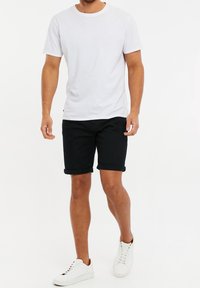 Homme portant un t-shirt blanc uni, un short noir retroussé et des baskets blanches marchant sur un fond blanc.
