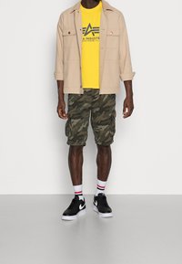 Bežová sako s knoflíky přes žluté tričko s potiskem, doplněné zelenými maskáčovými cargo šortkami a černými teniskami Nike s bílou podrážkou.