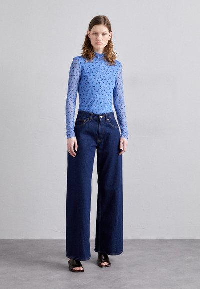 Maglietta blu a maniche lunghe con motivo floreale, abbinata a pantaloni in denim a vita alta e gamba larga. La modella indossa tacchi aperti neri.