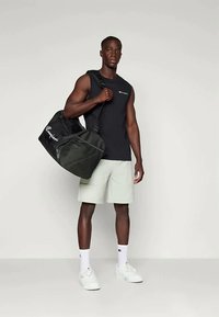 Musta urheilureppu logolla, teksturoitua kangasta, säädettävä olkahihna, henkilön päällä, joka pukeutuu ilmanhihaiseen paitaan ja vaaleisiin shortsit.