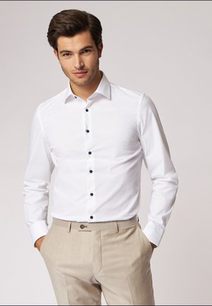 Weißes Langarmhemd mit Button-Down-Kragen und navyfarbenen Knöpfen. Kombiniert mit hellbeigen Hosen, die eine glatte Stofftextur aufweisen.
