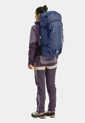 Blaue Wanderrucksack aus strukturiertem Stoff, mit mehreren Riemen und Reißverschlusstaschen, getragen über einem lila und schwarzen Kletteroutfit mit Akzentfarben.