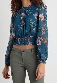 Teal blomsterbluse med lange gennemsigtige ærmer, smocket talje og lyserøde og hvide blomster mønstre. Matchet med olivengrønne bukser.