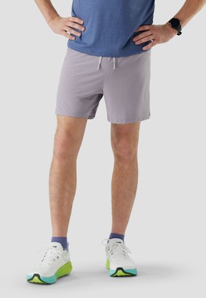 Mann trägt hellviolette Shorts, blaues Hemd, weiße und grüne Laufschuhe, steht mit den Händen in die Hüften gestützt vor grauem Hintergrund.