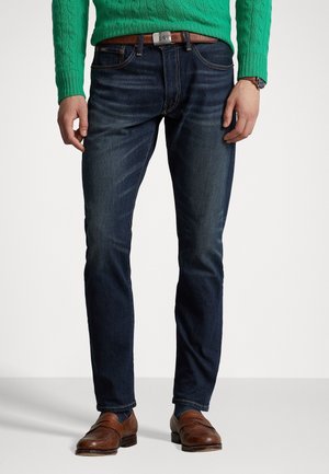 Hombre con vaqueros ajustados azul oscuro, cinturón de cuero marrón, mocasines de cuero marrón y un suéter verde de punto tipo trenza.