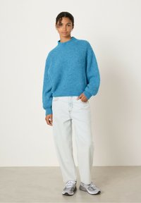 Mėlynas, tekstūruotas megztinis su aukštu kaklu ir plačiais rankovėmis, derinamas su šviesiai mėlynomis džinsinėmis kelnėmis ir pilkais sportiniais bateliais.