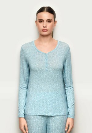 Donna che indossa una maglia henley a maniche lunghe azzurro chiaro e pantaloni abbinati con una discreta stampa leopardata, in piedi davanti a uno sfondo semplice.