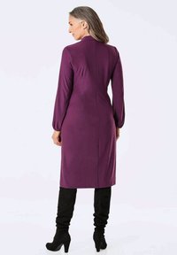 Lila, langärmliges Kleid mit gerader Silhouette und Rückennahtdetails, kombiniert mit schwarzen, oberschenkelhohen Stiefeln, vor einem hellen Hintergrund.